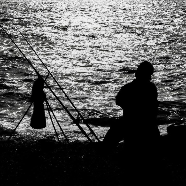 Ein Angler schaut aufs Meer. Die Angel steht Links neben ihm. Das Bild ist schwarz weiß